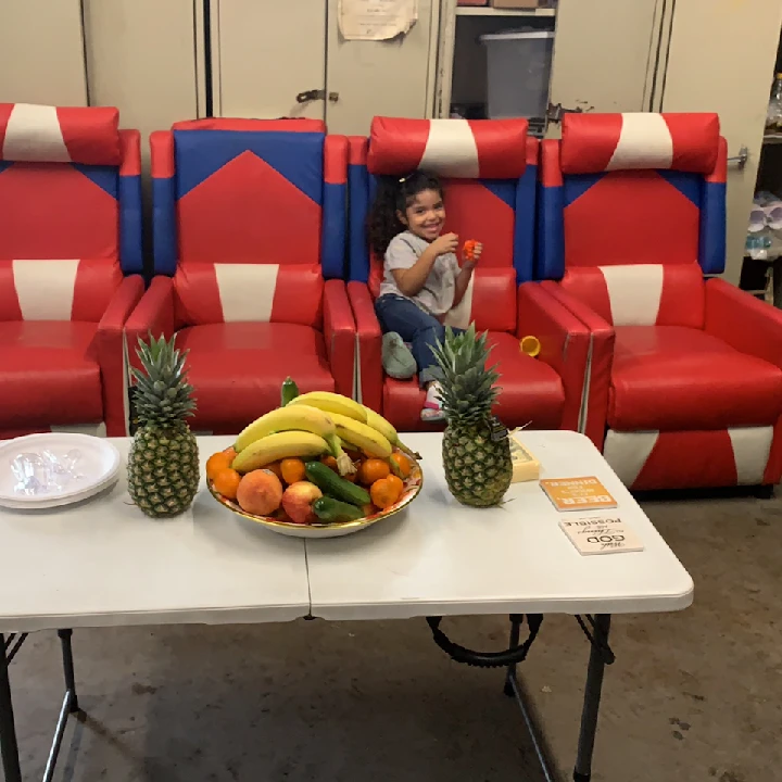 Child sitting on red chairs with fruit on a table.