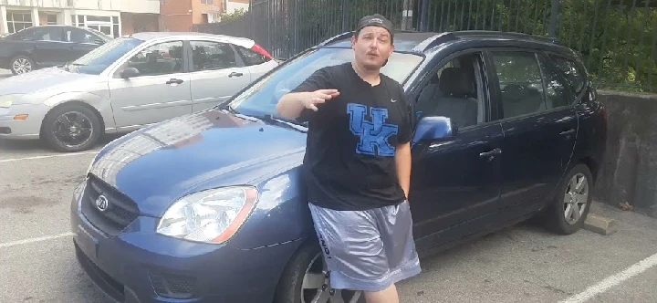 Man gesturing beside a blue car in a parking lot.