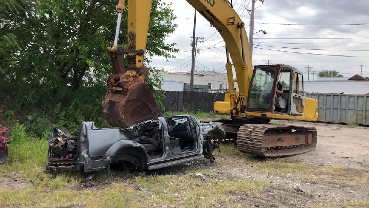 Excavator lifting a crushed vehicle in a junkyard.