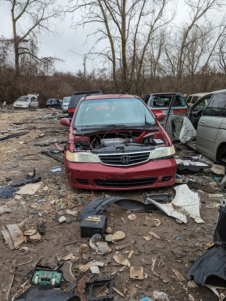 Rusty red car in a junkyard among scattered debris.