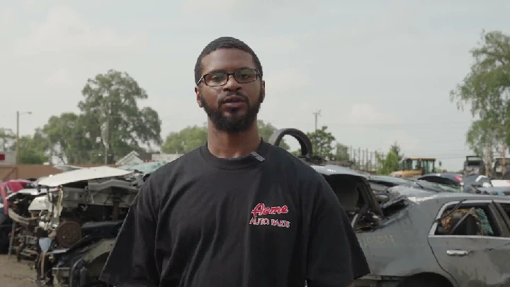 Man in Acme Auto Parts shirt at scrapyard.