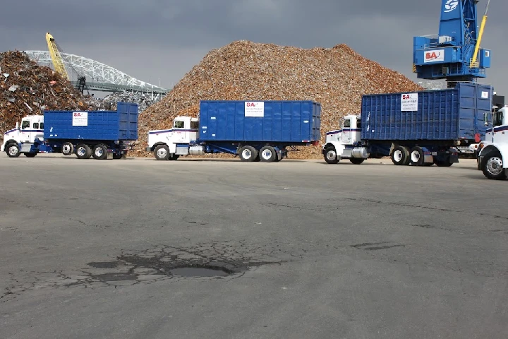 Blue trucks lined up near a large scrap metal pile.