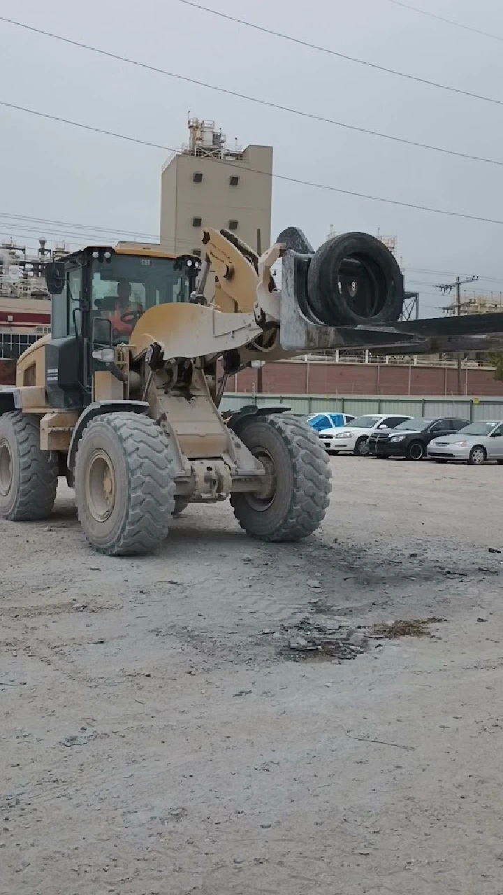 Heavy machinery lifting a tire in a gravel lot.